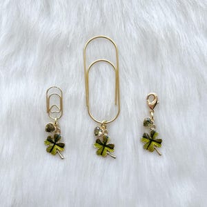 May include: Three gold-colored paper clips with decorative charms. Each charm features a green and yellow four-leaf clover design with a small, round, green gem. The paper clips are arranged on a white, fluffy surface.