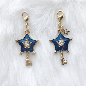 May include: Two gold-toned star charms with blue enamel and crystal accents. Each charm features a star shape with a key design at the bottom and a small star charm. The charms are attached to a gold clasp.