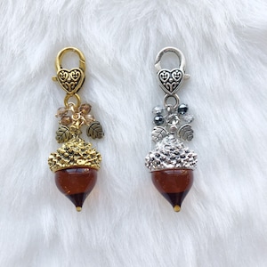 May include: Two acorn-shaped charms with lobster clasps. One charm has a gold-tone finish with brown beads and leaves, the other has a silver-tone finish with gray beads. Both charms have a brown, translucent acorn body.