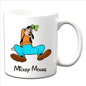 May include: White ceramic mug featuring a cartoon illustration of Goofy in an orange shirt, blue pants, and brown shoes. The mug has a black "Mickey Mouse" text at the bottom. The mug has a white handle.