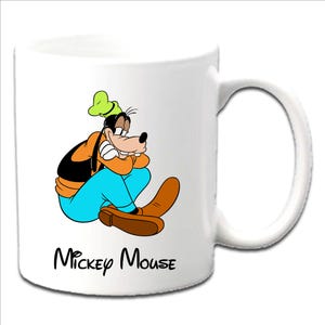May include: White ceramic mug featuring a colorful illustration of Goofy from Disney. Goofy is depicted in his classic outfit, with the text "Mickey Mouse" below the character. The mug has a white handle.