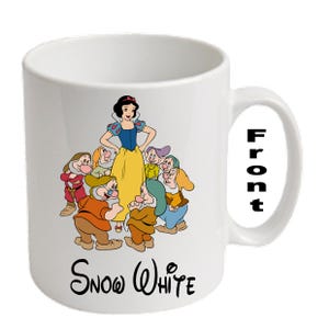 Op de afbeelding: Witte keramische mok met een kleurrijke illustratie van Sneeuwwitje en de Zeven Dwergen. De mok heeft een gebogen handvat en het woord "Snow White" in script onderaan.