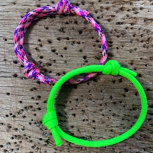 May include: Two friendship bracelets made of colorful paracord. One bracelet is pink, purple, and white, and the other is bright green.