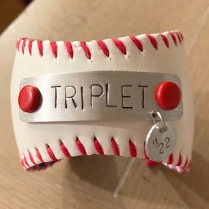 May include: White leather baseball cuff bracelet with red stitching. The bracelet has a silver metal plate with the word "TRIPLET" engraved on it. There is a small silver charm with the number "#22" on it.