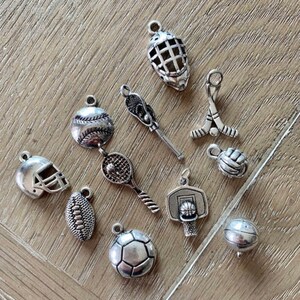 May include: A collection of silver-toned sports charms, including a baseball, basketball, football, soccer ball, hockey stick and puck, lacrosse stick, tennis racket, and a hockey goalie mask.