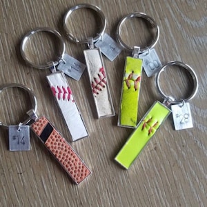 May include: Five keychains with different baseball-themed designs. Each keychain features a small metal tag with a number, such as #14, #4, #00, and #28. The keychains are made from real baseballs and have a variety of colors, including white, brown, yellow, and green.