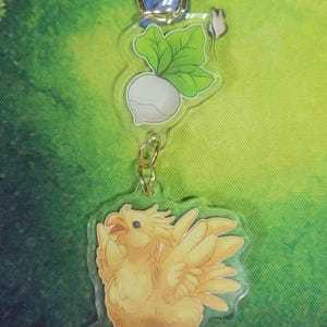 May include: A clear acrylic keychain featuring a yellow Chocobo bird charm, a white radish charm, and a gold star-shaped clasp. The Chocobo has detailed feathers and a beak. The radish has green leaves.