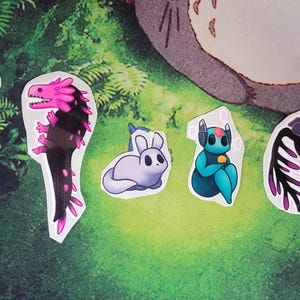 Op de afbeelding: Een verzameling grillige stickers met verschillende fantasie wezens. De stickers bevatten een bruine eland met een klein wezen op zijn gewei, een roze en zwarte draak, een paars konijn, een turquoise robotachtig figuur en een zwart-roze vogel.