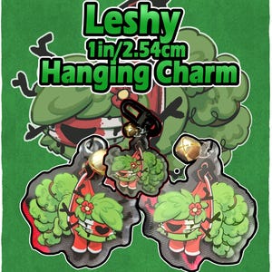 May include: A green background features a cartoon-style charm with the text "Leshy Hanging Charm." The charm is 1 inch (2.54 cm) and depicts a character with green foliage, a red body, and a flower. The charm has a black clip and gold bells.