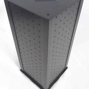May include: A black rotating display stand with a square base and a perforated surface. The stand is designed for displaying small items, such as jewelry or accessories.
