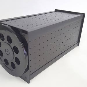 May include: Black storage box with a round, perforated lid. The lid has a series of holes around the edge.