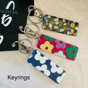 Peut inclure: Trois porte-clés avec des motifs différents. Les porte-clés sont attachés à des anneaux de clés argentés. Les motifs sont un motif géométrique gris foncé et beige, un motif floral rose et violet et un motif floral bleu et blanc. Les porte-clés sont étiquetés "Porte-clés".