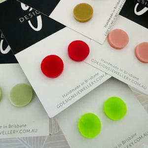 Pode incluir: Um conjunto de seis pares de brincos redondos de botão em várias cores, incluindo vermelho, verde, rosa e dourado. Os brincos são exibidos em cartões brancos com o texto "Handmade in Brisbane GDESIGNSJEWELLERY.COM.AU".