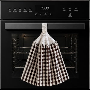 May include: A hanging kitchen towel featuring a brown and white chequered pattern with a white crocheted top section. The towel is secured with a single white button at the top, designed to hang from a handle. The fabric has a textured, waffle-like weave.