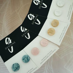 May include: Assortment of six pairs of earrings displayed on cards. The earrings are made of different materials, including shell, stone, and wood. The cards are black with white text that reads "G Designs".