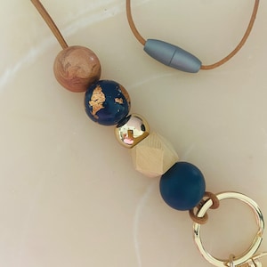 May include: A brown leather lanyard with a gold metal clasp and a key ring. The lanyard has five beads: two dark blue with gold flecks, one light brown, one gold, and one dark blue.