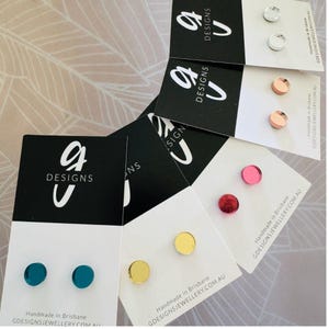 May include: Assortment of small, round, mirrored earrings in various colors, including gold, silver, teal, red, pink, and copper. The earrings are displayed on white cards with the text "G Designs" and "Handmade in Brisbane" printed on them.