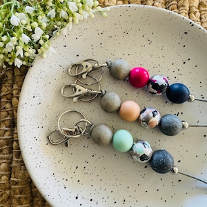 Op de afbeelding: Drie sleutelhangers met zilveren sleutelringen en gekleurde kralen. De kralen zijn grijs, roze, lichtblauw en donkerblauw met witte accenten. De kralen zijn geregen aan een zilveren ketting.