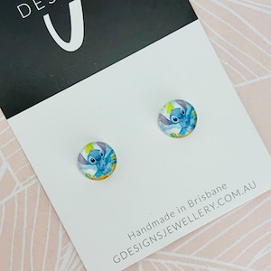 Puede incluir: Un par de pendientes de bot&oacute;n redondos con el personaje Stitch de la pel&iacute;cula Lilo & Stitch. Los pendientes est&aacute;n colocados sobre una tarjeta blanca con el texto "GDESIGNSJEWELLERY.COM.AU" y "Handmade in Brisbane".