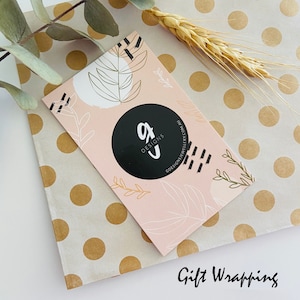 Puede incluir: Una tarjeta de visita con el logo "gJ Designs" sobre un fondo rosa con dise&ntilde;os de hojas y abstractos. La tarjeta est&aacute; sobre papel de regalo de lunares, con espigas de trigo y eucalipto.