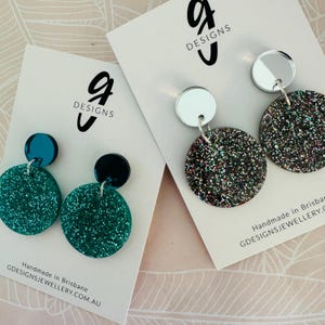 May include: Two pairs of earrings with glitter and mirror accents. The earrings are on white cards with the text "G Designs" on them. The cards also have the text "Handmade in Brisbane GDESIGNSJEWELLERY.COM.AU".