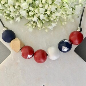 Può includere: Una collana di perline rosse, bianche e blu con accenti argentati. La collana è realizzata con perline di legno e ha un cordoncino nero.