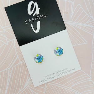 Puede incluir: Un par de pendientes de bot&oacute;n redondos con un dise&ntilde;o de personaje de dibujos animados azul. Los pendientes se muestran en una tarjeta blanca con el texto "G DESIGNS" y "GDESIGNSJEWELLERY.COM.AU". El fondo es un patr&oacute;n floral rosa claro.
