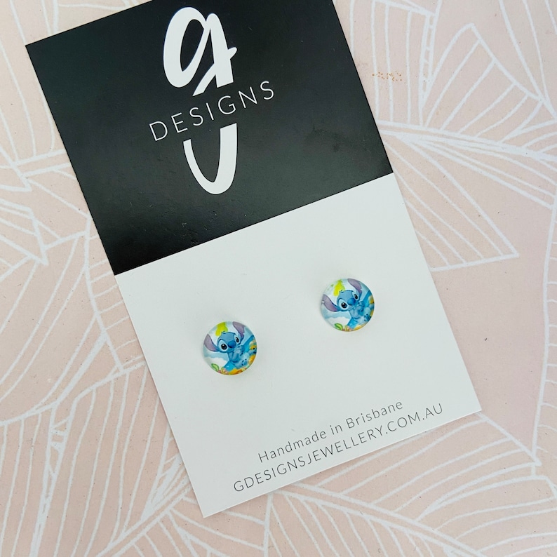 Puede incluir: Un par de pendientes de bot&oacute;n redondos con un dise&ntilde;o de personaje de dibujos animados azul. Los pendientes se muestran en una tarjeta blanca con el texto "G DESIGNS" y "GDESIGNSJEWELLERY.COM.AU". El fondo es un patr&oacute;n floral rosa claro.