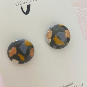 May include: A pair of round stud earrings with a black, white, gold, and light blue abstract pattern.