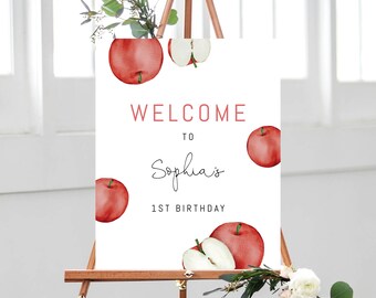 Op de afbeelding: Welkom bij Sophia's 1e verjaardag bord met rode appel illustraties. Het bord is wit met zwarte tekst en staat op een houten ezel.
