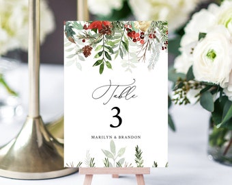 Pode incluir: Um cartão branco com número de mesa com detalhes florais em aquarela. O cartão diz "Table 3" em escrita preta e inclui os nomes "Marilyn & Brandon". A borda superior apresenta folhas verdes, bagas vermelhas e pinhas. O cartão é exibido em um pequeno cavalete de madeira.