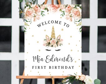 May include: Welcome to Mia Edmond's First Birthday sign with a gold glitter unicorn and floral design. The sign has a white background with gold glitter stars and confetti.