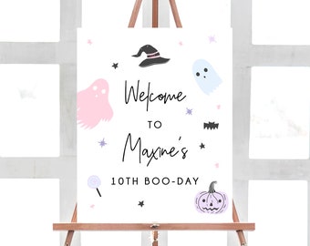 Puede incluir: Un cartel blanco con un fantasma rosa y azul, un sombrero de bruja negro y una calabaza morada. El cartel dice "Bienvenido a Maxine's 10th Boo-Day".