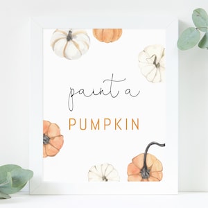 Puede incluir: Un estampado enmarcado con calabazas de acuarela en tonos naranja y blanco sobre un fondo blanco. El texto "paint a PUMPKIN" está escrito en escritura negra y naranja. Ramas de eucalipto en el fondo.