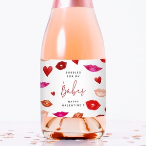 Puede incluir: Una botella de champán rosa con una etiqueta blanca que dice "Bubbles for my Babes Happy Galentine's". La etiqueta está decorada con labios y corazones rojos y rosas.