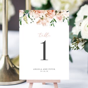 Puede incluir: Una tarjeta de número de mesa blanca con un diseño floral en la parte superior. La tarjeta dice "mesa 1" en texto negro y "Angela y Peter 01.19.19" en texto negro más pequeño.