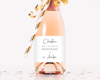 Peut inclure: Une mini bouteille de champagne avec une étiquette blanche et du texte noir. L'étiquette indique "Christine, Veux-tu être ma demoiselle d'honneur ?  xo. Amber" avec un cœur rose.