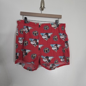 May include: Red boxer shorts featuring an all-over print of Mickey Mouse heads, winged emblems, and circular logos with the letter M. The shorts have an elastic waistband and are displayed on a wooden hanger against a plain white wall.