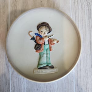 Könnte beinhalten: Eine cremefarbene dekorative Platte mit einer 3D-Figur eines Geige spielenden Jungen. Der Junge trägt einen braunen Hut, eine orangefarbene Jacke, grüne Hosen und trägt einen Regenschirm. Die Platte hat einen erhöhten Rand und den Text "M.I. Hummel" an der Basis.