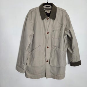 May include: A beige utility jacket featuring a dark olive green corduroy collar and matching cuffs. The jacket includes a front button closure with brown buttons, two large lower patch pockets, and a single zippered chest pocket on the left side.