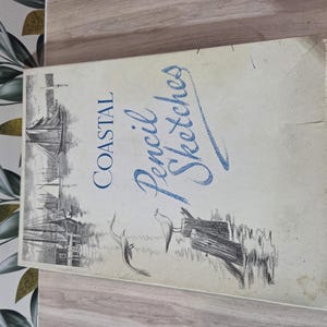 May include: A rectangular book titled Coastal Pencil Sketches with a cream-colored cover. The front features a black and white illustration of a sailboat and two birds perched on wooden posts. The title is printed in blue and black text.