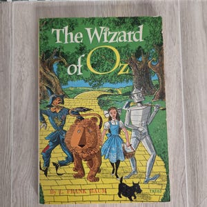 May include: A paperback book titled The Wizard of Oz by L. Frank Baum. The cover features a colorful illustration of Dorothy, the Scarecrow, the Tin Woodman, the Cowardly Lion, and a small black dog walking along a yellow brick road in a forest.