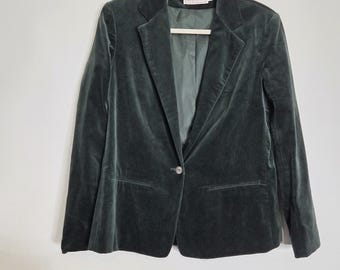 May include: A dark forest green velvet blazer featuring a single-button closure, notched lapels, and two front welt pockets. The jacket is fully lined in a matching green fabric and includes a white brand label at the inner neckline.