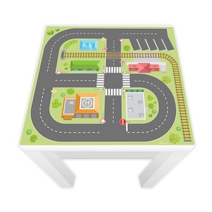 Puede incluir: Una mesa blanca con una alfombra de juego verde que presenta un sistema de carreteras con un helipuerto, vías de tren, estacionamiento y una señal de alto. La alfombra de juego está diseñada para coches y trenes de juguete.