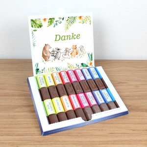 Pode incluir: Uma caixa de barras de chocolate ao leite sortidas com embalagens coloridas que apresentam várias mensagens. A caixa está aberta e a aba superior tem um cartão com uma mensagem "Danke" verde e uma ilustração de desenhos animados de animais.