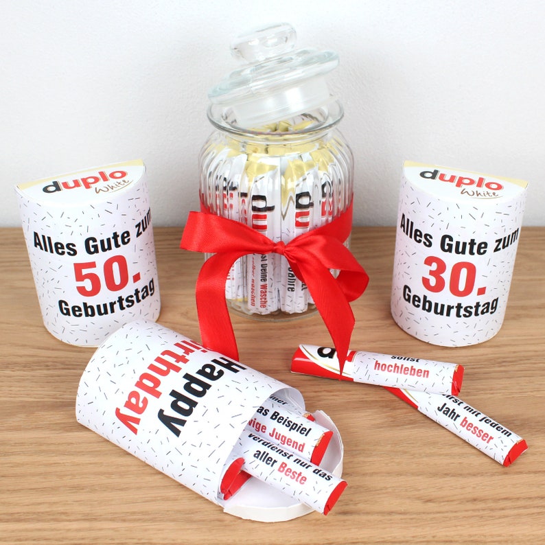 May include: A glass jar filled with white chocolate bars wrapped in red and white paper with a red ribbon tied around the jar. The jar is surrounded by three white cardboard boxes with red and black text. The boxes have the text "duplo White" and "Alles Gute zum 50. Geburtstag" and "Alles Gute zum 30. Geburtstag". There are also three white chocolate bars with red and black text on the wrappers.
