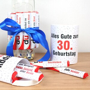 May include: A white mug with black polka dots and the text "Alles Gute zum 30. Geburtstag" in red and black. The mug is surrounded by chocolate bars wrapped in red and white paper with various sayings in German.