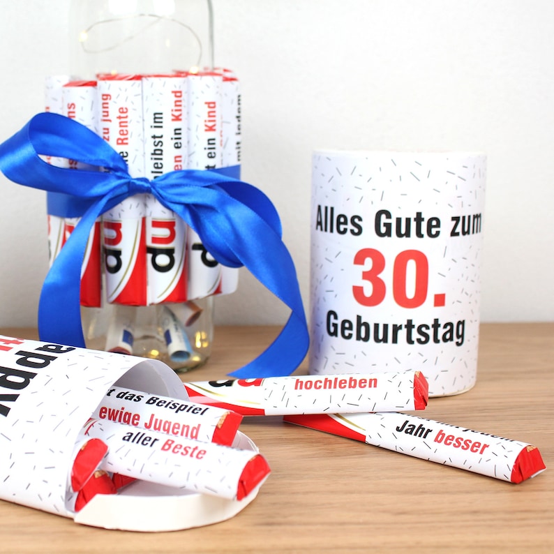 May include: A white mug with black polka dots and the text "Alles Gute zum 30. Geburtstag" in red and black. The mug is surrounded by chocolate bars wrapped in red and white paper with various sayings in German.