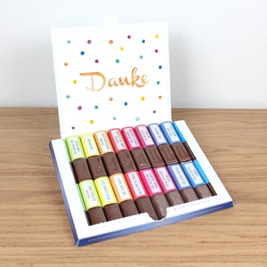 Puede incluir: Una caja de chocolates con leche surtidos con envoltorios coloridos y mensajes en alemán. La caja está abierta y los chocolates están dispuestos en filas. La parte superior de la caja tiene una tarjeta blanca con confeti de colores y la palabra "Danke" escrita en naranja.