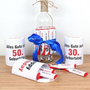 May include: A birthday gift idea featuring a glass bottle filled with chocolate bars wrapped in red and white paper. The bottle is decorated with a blue ribbon and a string of lights. The bottle is surrounded by two white cardboard tubes with the text "Alles Gute zum 50. Geburtstag" and "Alles Gute zum 30. Geburtstag".
